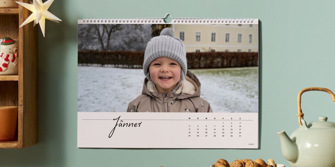 Ein fröhliches Kind mit einer Mütze im Schnee lächelt vor einem Wandkalender.