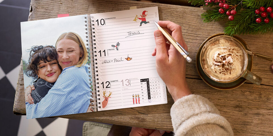 Offener CEWE Fotokalender mit Terminseite und Foto zweier Freundinnen auf einem Holztisch neben Tasse und Tannenzweig