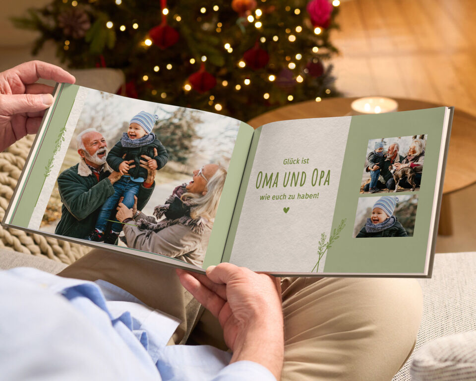 Person betrachtet ein CEWE FOTOBUCH mit Familienfotos und dem Schriftzug „Glück ist OMA UND OPA wie euch zu haben!“ vor einem Weihnachtsbaum