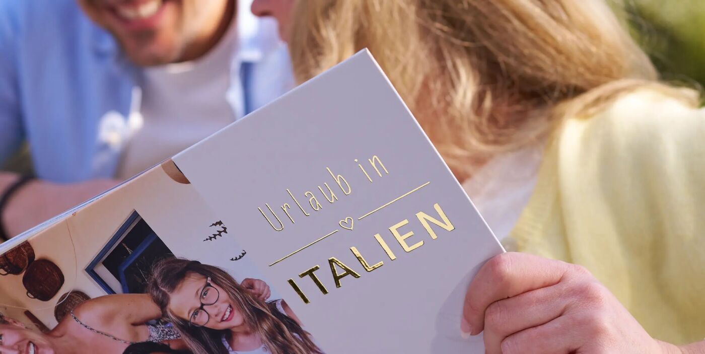 Eine Person hält ein CEWE FOTOBUCH mit dem Titel „Urlaub in Italien“ mit Goldveredelung in der Hand.