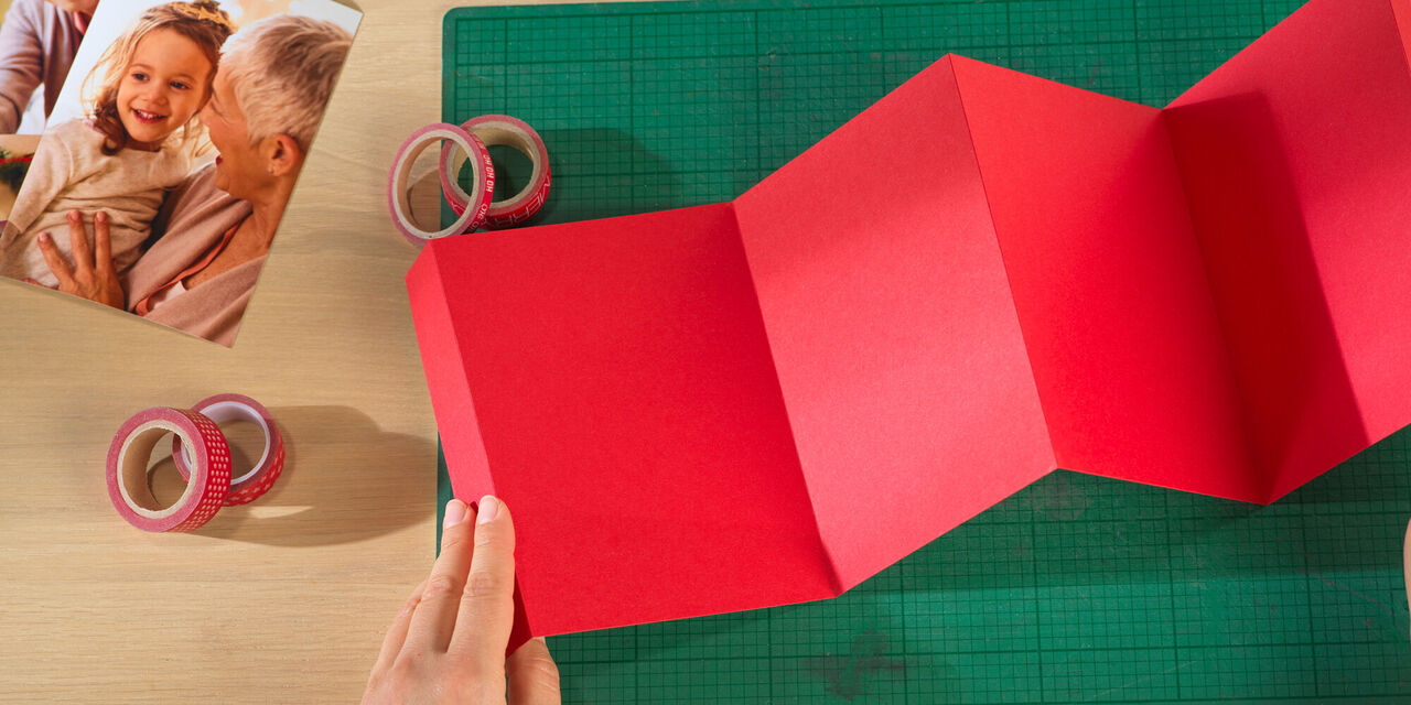 Jemand hält eine rote Faltkarte auf einem Tisch, umgeben von buntem Klebeband und einem Foto.