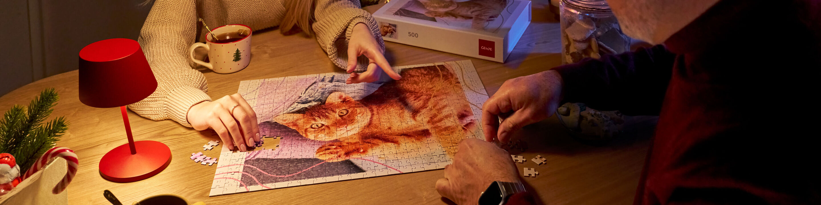 Zwei Personen setzen gemeinsam ein Foto-Puzzle mit Katzenfoto zusammen, daneben stehen eine rote Tischlampe und Weihnachtsdekoration.