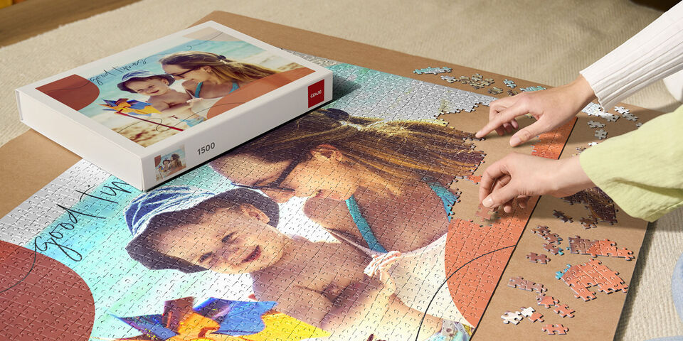 Zwei Personen, eine Frau und ein Kind, legen zusammen ein Puzzle mit einem bunten Bild darauf. Der Puzzlekarton liegt daneben.