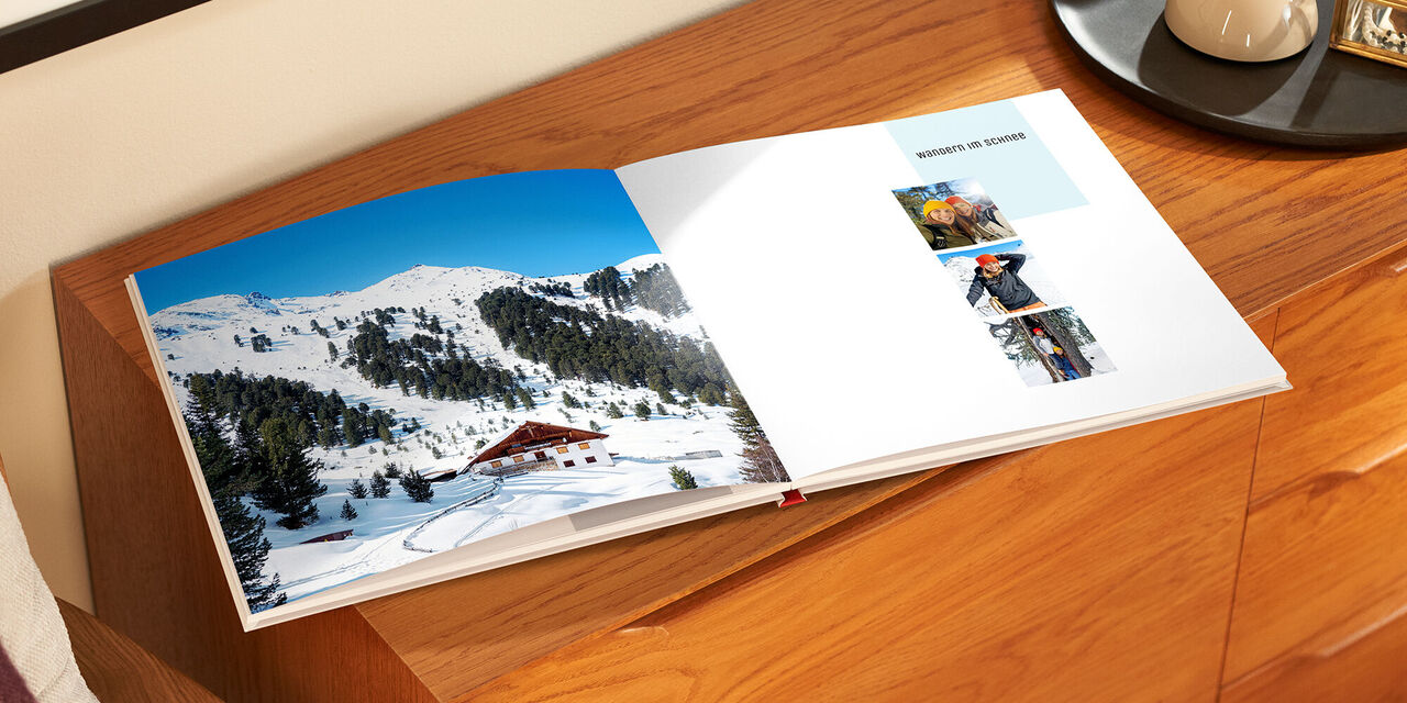 Auf einem Holzschrank liegt das aufgeklappte Fotobuch. Links sieht man ein vollflächiges Foto eines Gasthauses vor einer verschneiten Bergkette. Auf der rechten Seite sind drei quadratische Bilder angeordnet, die jeweils zwei Frauen zeigen.