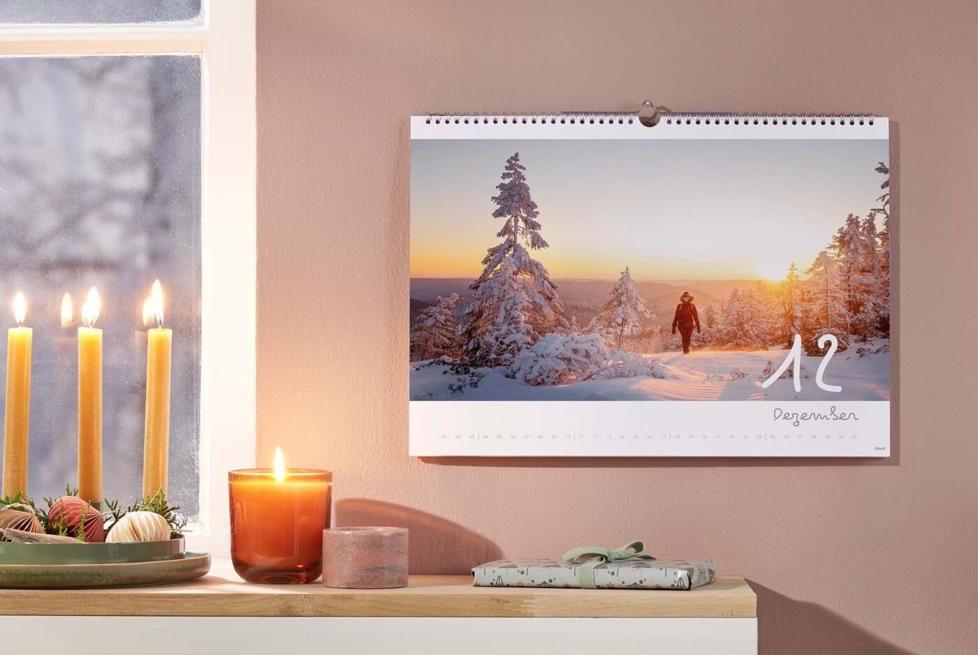 Ein schöner Wandkalender für den Dezember zeigt eine Person, die durch schneebedeckte Bäume in der Winterlandschaft spaziert. Das warme Licht der Abendsonne strahlt über die Szene.